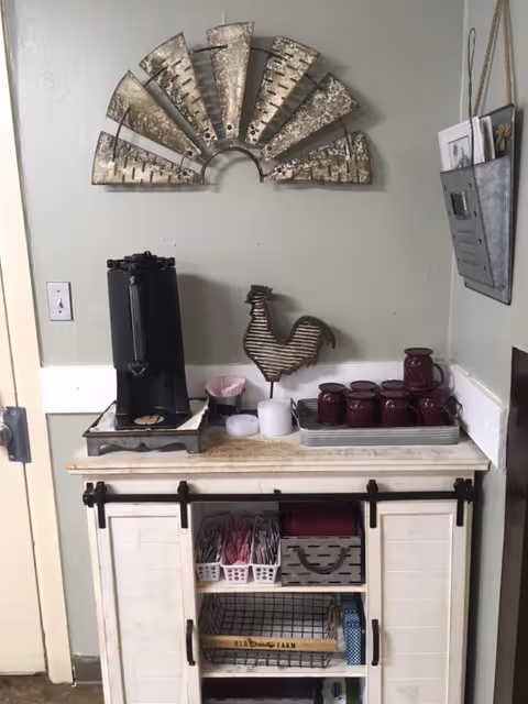A small beverage station with a black coffee dispenser on a white wooden cabinet. The cabinet has sliding barn-style doors and open shelves holding baskets with coffee supplies like sugar packets and stirrers. On top of the cabinet, there is a metal rooster decoration, several maroon mugs, and a metal tray. Above the station, a decorative metal wall art piece resembling a fan is mounted on a light gray wall. To the right, a metal wall organizer holds papers and envelopes.