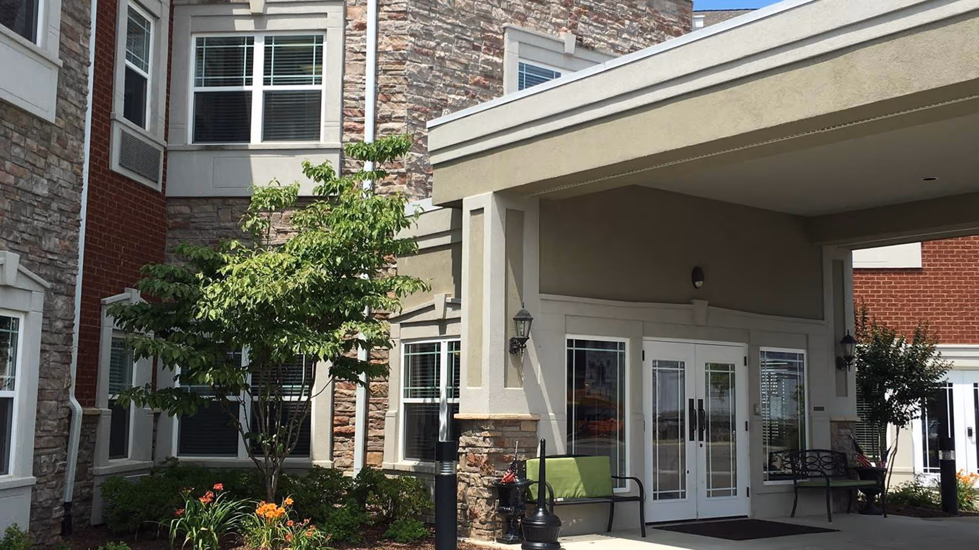 Entrance area of Maristone of Providence facility showing a covered drop-off with double glass doors, stone and brick exterior walls, green benches, small trees, and landscaped flower beds.