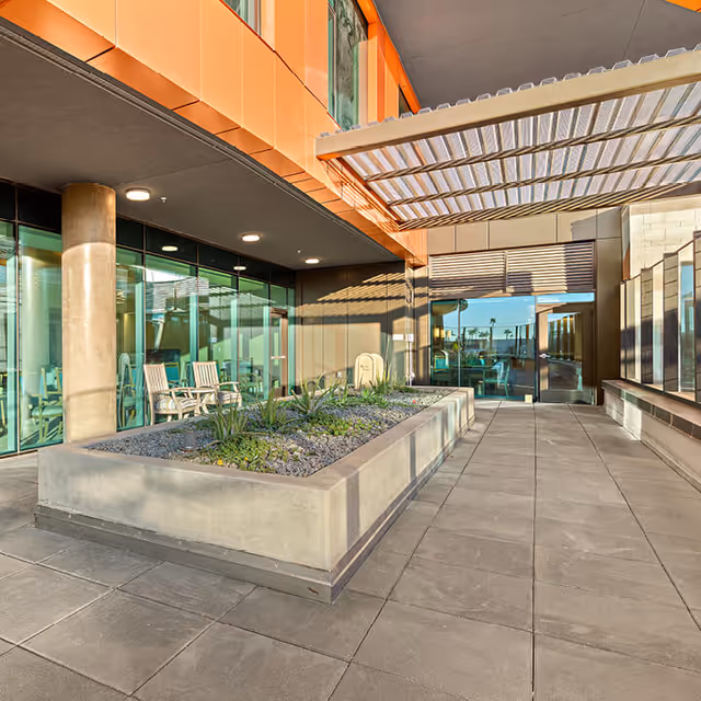 Outdoor patio area at Mirabella at ASU featuring a large rectangular planter with green plants and rocks, surrounded by tiled flooring. There are glass walls and doors leading inside the building, with a shaded pergola overhead and modern architectural elements including orange paneling and concrete columns.