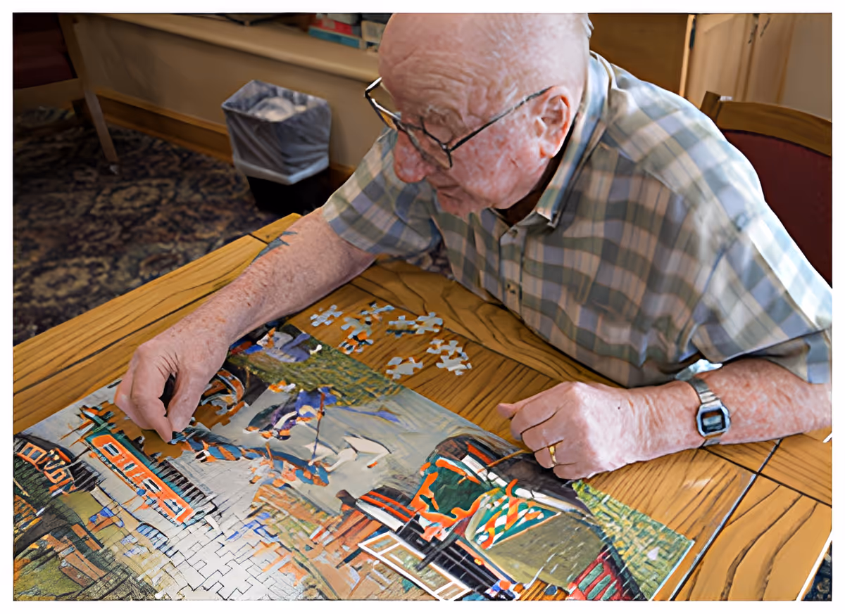 An elderly man wearing glasses and a plaid shirt is sitting at a wooden table working on assembling a colorful jigsaw puzzle. The room has a patterned carpet and a trash bin is visible in the background.