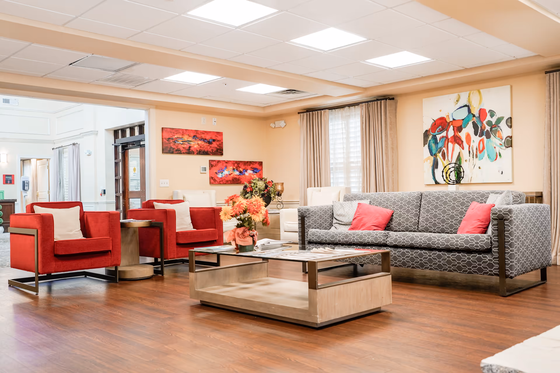 A bright and spacious living room area in a senior living facility featuring a gray patterned sofa with red and gray cushions, two red armchairs with white pillows, a wooden coffee table with a glass top, and colorful abstract paintings on the walls. The room has wood flooring, beige walls, and large windows with beige curtains allowing natural light to enter.