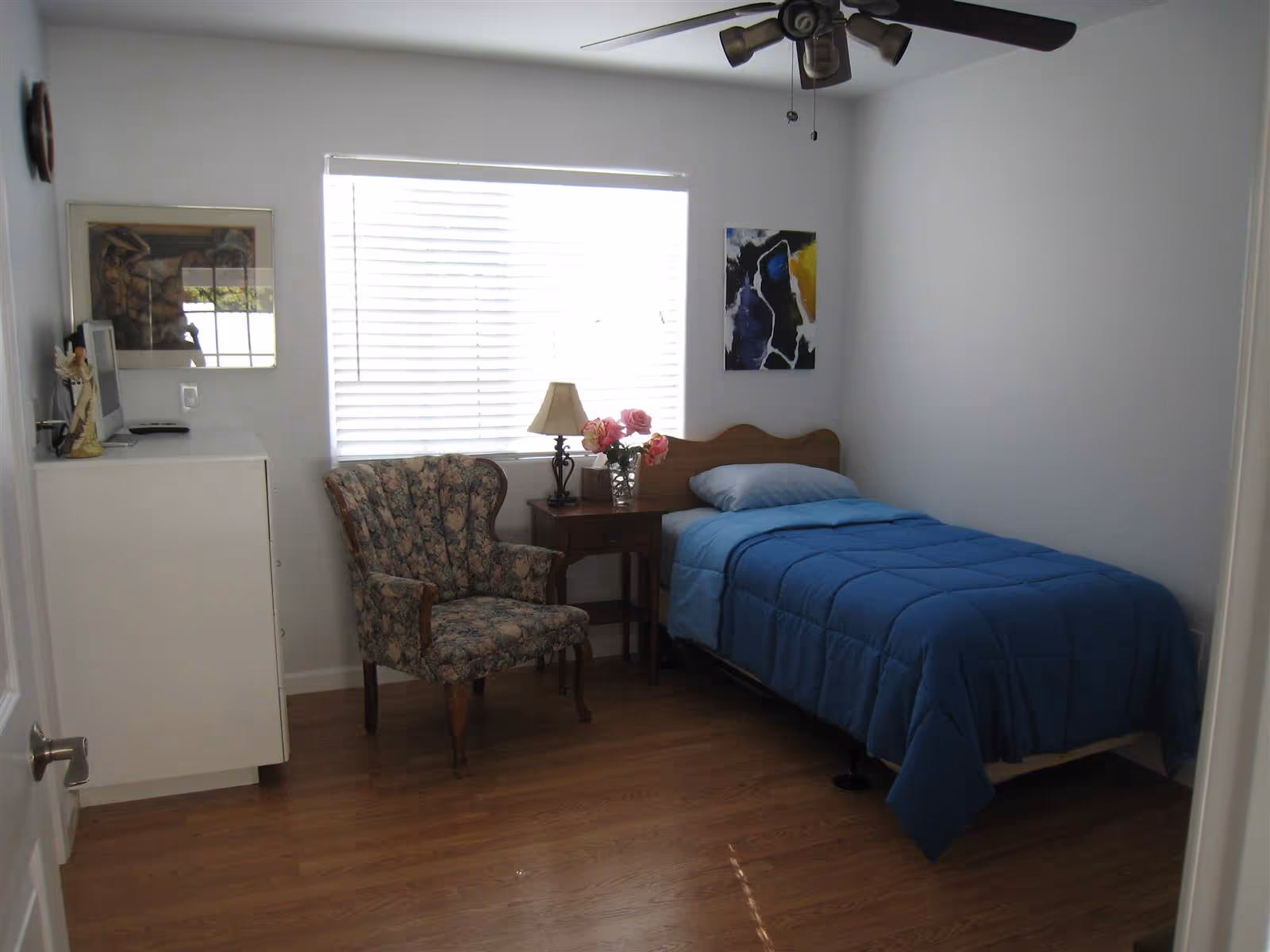 A small bedroom with a single bed covered in a blue comforter and a light blue pillow. Next to the bed is a wooden nightstand with a lamp and a vase of pink flowers. A floral upholstered armchair is positioned near a white dresser with a small TV on top. The room has wooden flooring, white walls, a ceiling fan with lights, and a window with closed blinds letting in natural light. Two framed artworks hang on the walls.