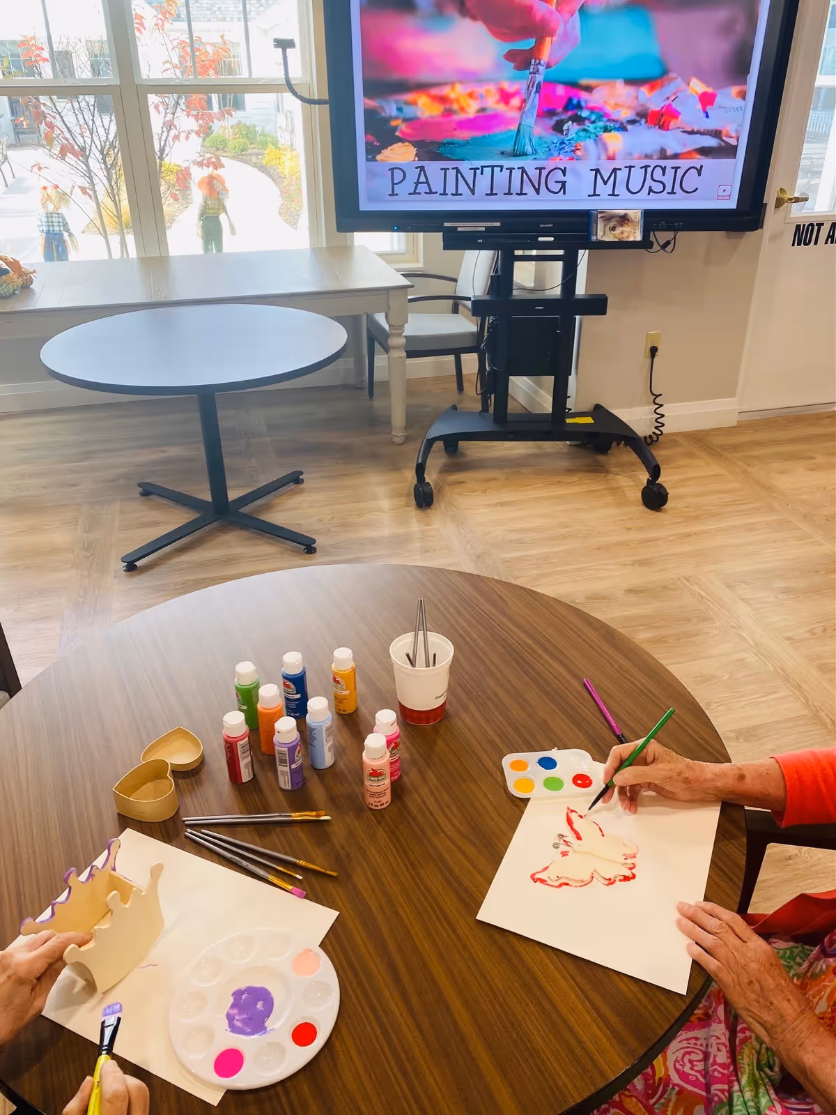 Two elderly individuals seated at a round wooden table engaged in painting activities. One person is painting a wooden crown with purple paint, while the other is painting a butterfly outline on paper with red paint. Various paint bottles, brushes, and palettes are arranged on the table. In the background, a large screen displays the words 'PAINTING MUSIC' with an image of a paintbrush and colorful paint. The room has large windows showing an outdoor path with autumn foliage.