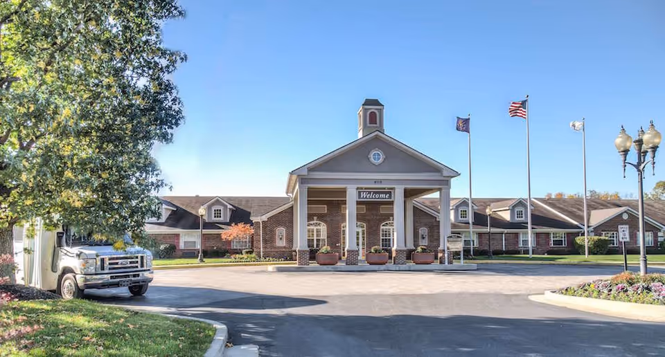 Front exterior view of a single-story brick building with a covered entrance supported by white columns. There are three flagpoles with flags, a street lamp, and a truck parked partially behind a tree on the left side. The sky is clear and blue.