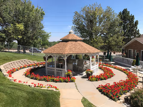 A well-maintained outdoor garden area featuring a white gazebo with a brown shingled roof surrounded by vibrant red and yellow flowers arranged in circular flower beds. There are green trees and a clear blue sky in the background, along with a brick building and outdoor seating with umbrellas.