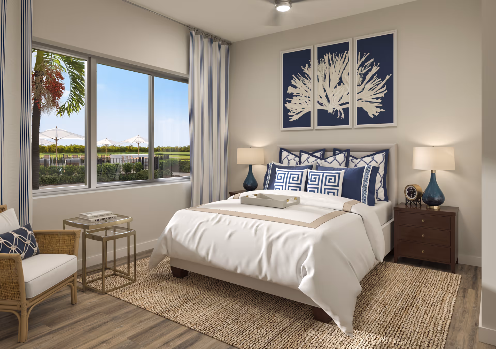 A bright and neatly decorated bedroom featuring a large bed with white and blue bedding, two bedside tables with matching blue lamps, a beige woven rug, a comfortable chair with a blue cushion, and a large window showing an outdoor view with umbrellas and greenery. Above the bed, there are three framed artworks depicting white coral on a blue background.