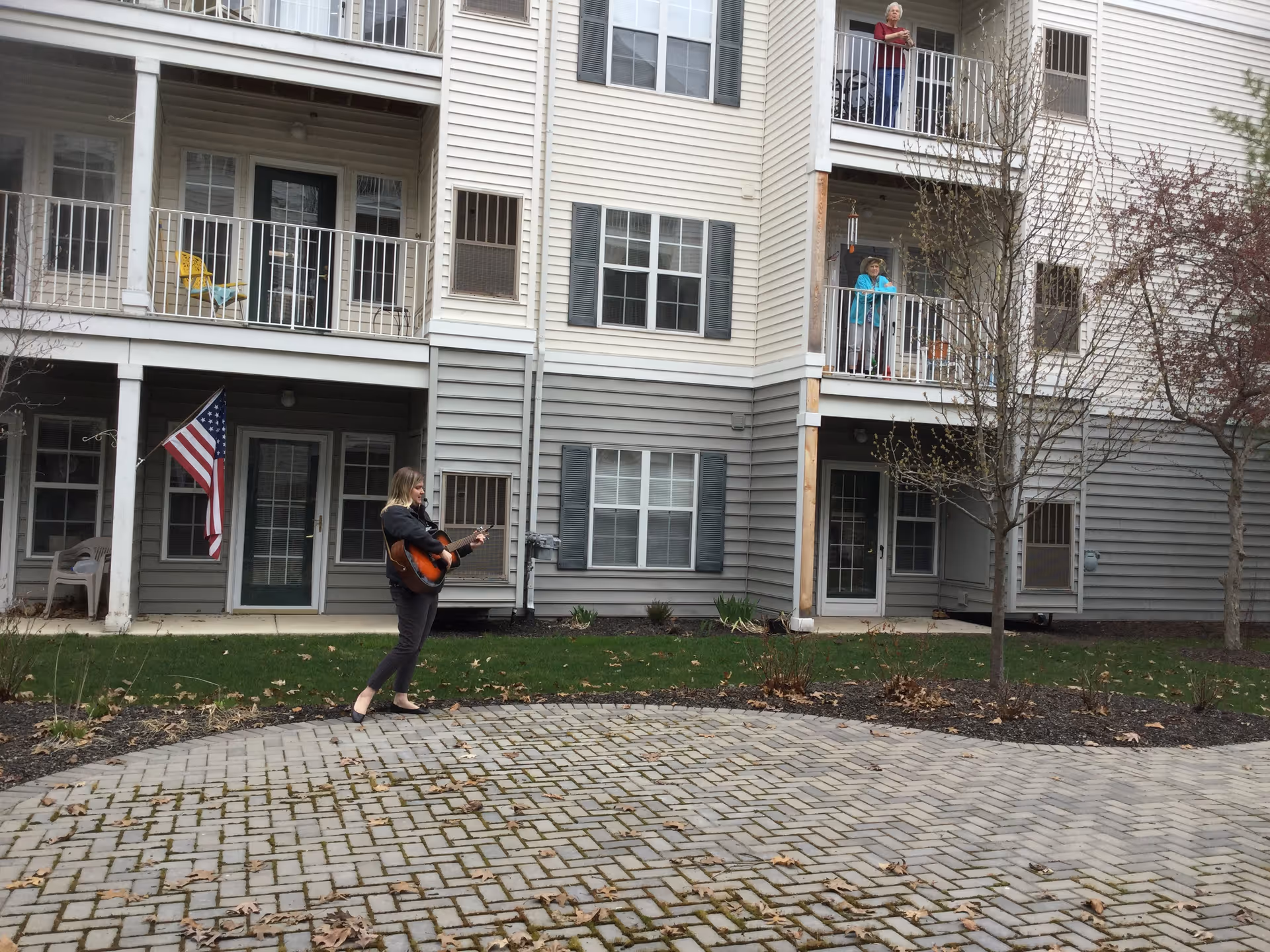 A woman playing guitar outside in front of a multi-story residential building with balconies. Two elderly women are standing on separate balconies watching her. The building has beige and gray siding with white railings and an American flag hanging near one of the doors.
