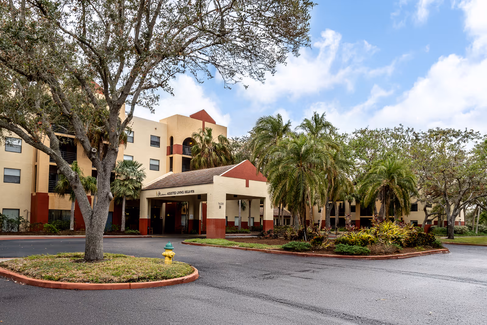 Front entrance of a multi-story assisted living building with a covered porte-cochere, palm trees, and a driveway.