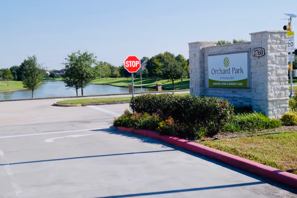 Entrance to Orchard Park at Victory Lakes Assisted & Senior Living facility with a stone sign displaying the facility name and services, a stop sign, a road, landscaped bushes, and a lake with trees in the background under a clear sky.