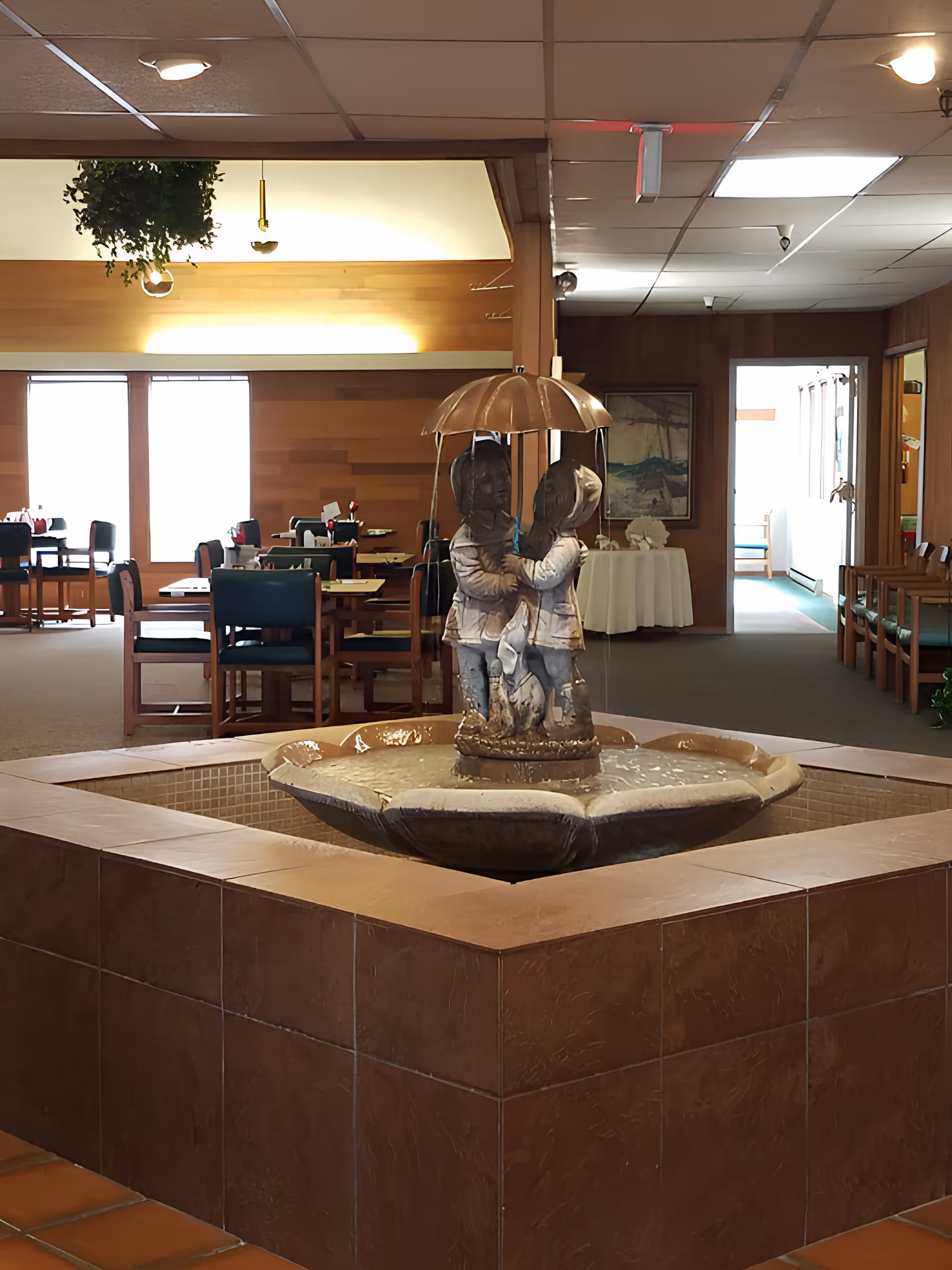 Indoor area of a retirement community featuring a central water fountain with a sculpture of two children holding an umbrella. Surrounding the fountain are dining tables and chairs, with wood-paneled walls and ceiling lights. A hallway and additional seating are visible in the background.