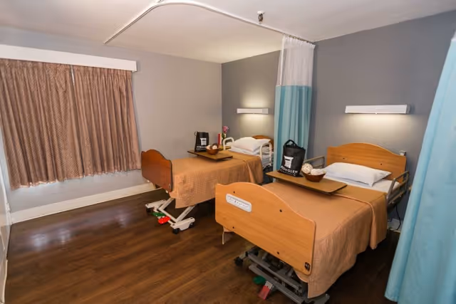 A room with two hospital-style beds with wooden headboards and footboards, each covered with a beige blanket. Each bed has a tray table with items on it, and there is a curtain divider between the beds. The room has wood flooring, a window with closed brown curtains, and soft wall lighting above each bed.