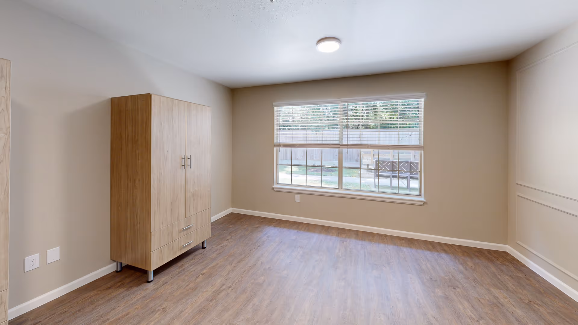 Empty bedroom with wood-look flooring, a wooden wardrobe, and a large window overlooking a fenced yard.