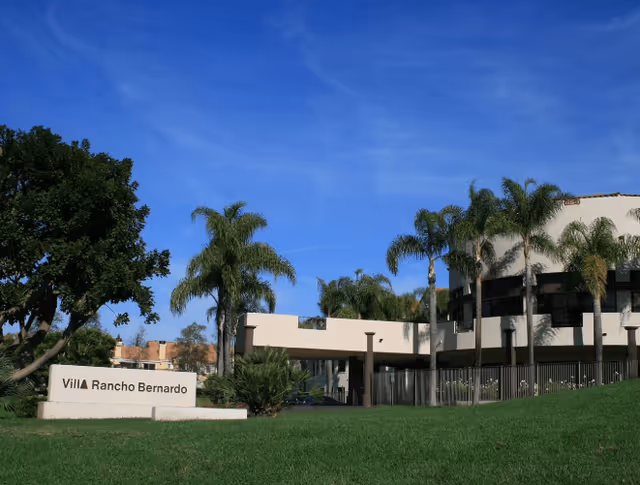 Exterior view of Villa Rancho Bernardo Care Center showing a modern building with palm trees and a green lawn under a clear blue sky. A sign with the facility name is visible on the left side.