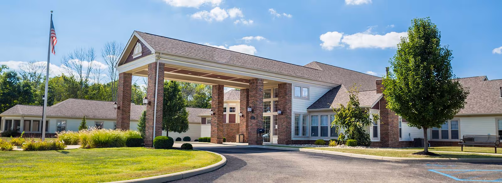 Exterior view of Altercare Zanesville facility showing a large covered entrance supported by brick columns, a well-maintained lawn, trees, and a flagpole with an American flag under a partly cloudy sky.