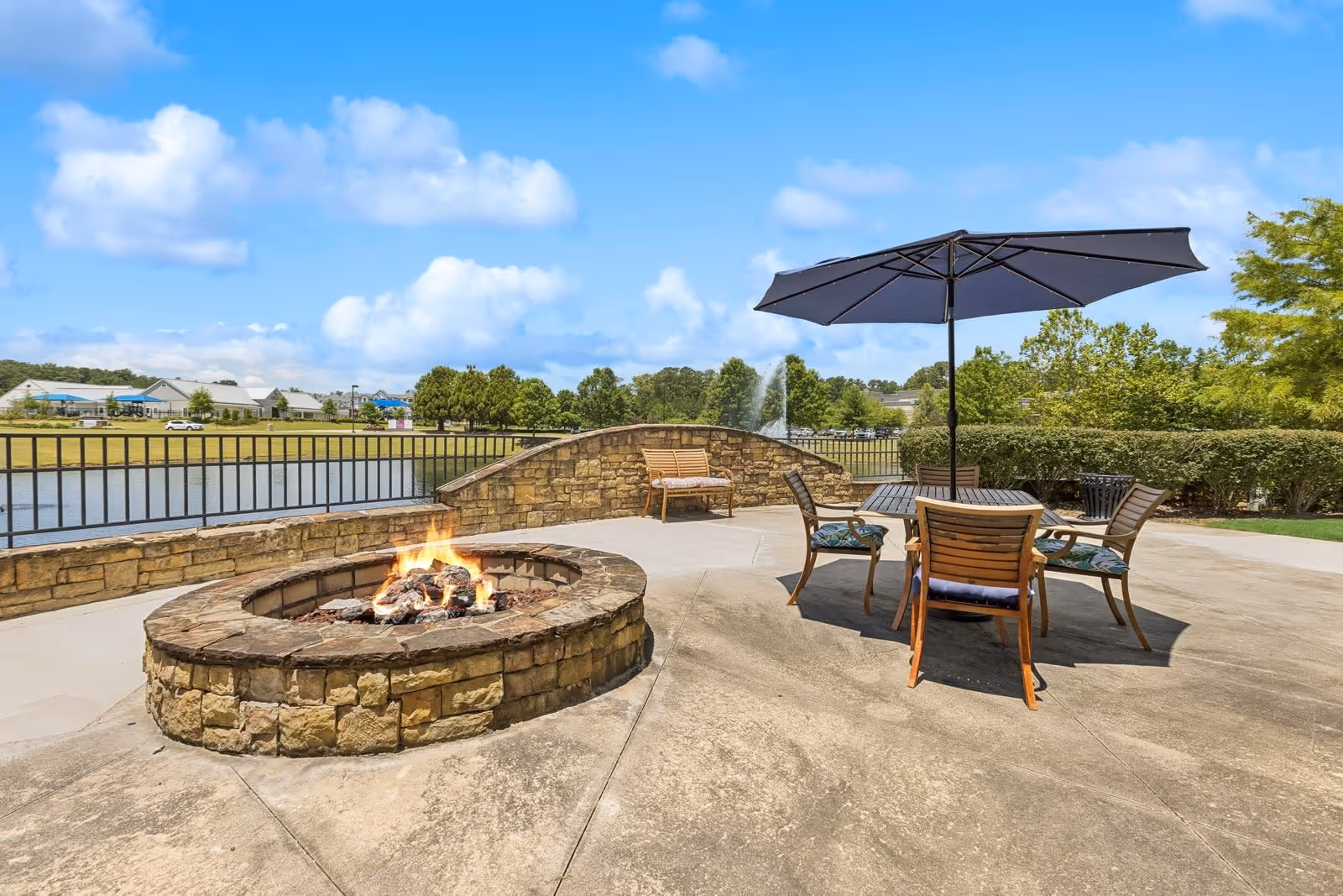 Outdoor patio area with a stone fire pit burning, surrounded by a concrete surface. There is a round table with four wooden chairs and a large black umbrella providing shade. In the background, there is a stone wall with a bench, a metal railing, a pond with a fountain, and trees under a blue sky with some clouds.