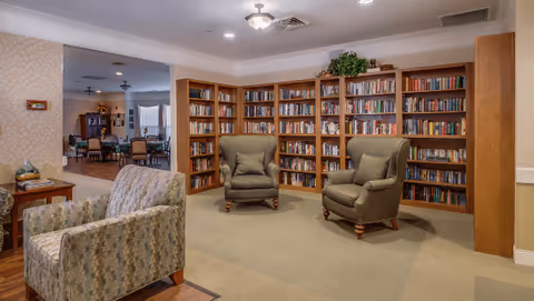 A cozy reading area in a senior living facility featuring three upholstered armchairs arranged around a carpeted floor, with a large wooden bookshelf filled with books against the wall. In the background, there is a dining area with tables and chairs, and soft lighting from ceiling fixtures.