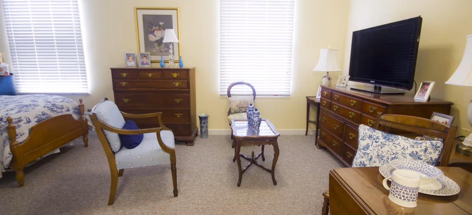 A cozy room with a wooden bed on the left, a cushioned armchair with a blue pillow, a wooden dresser with framed photos and a lamp, a small wooden table with decorative items, and a large wooden dresser with a flat-screen TV and lamps. The room has two windows with blinds and light-colored walls.