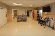 Interior view of a rehabilitation and healthcare center room with beige walls and floor. The room contains two blue chairs, a desk with a computer and office chair, a mounted flat-screen TV, a bookshelf, and medical equipment. There are doors leading to other rooms and a fire extinguisher mounted on the wall.