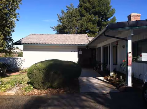 A single-story senior living facility front exterior with a walkway and trimmed shrubs.
