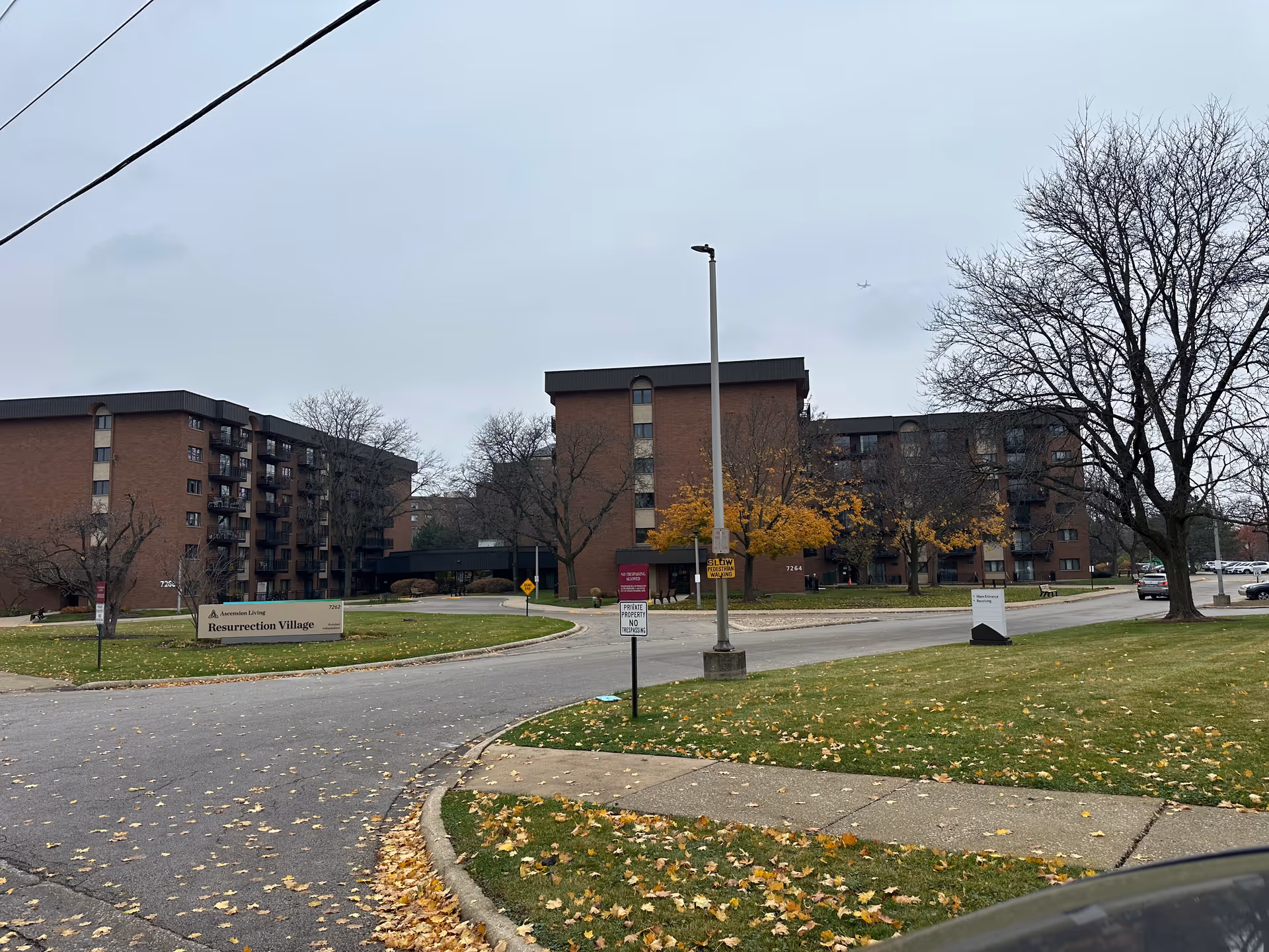 Large multi-story brick senior living complex with a circular driveway, a sign reading 'Resurrection Village', and trees with autumn leaves under an overcast sky.