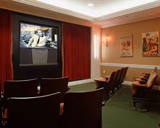 Small movie theater room with rows of brown cushioned chairs facing a large screen showing a black and white film. The room has green carpet, beige walls with white trim, red curtains framing the screen, and vintage movie posters on the walls.