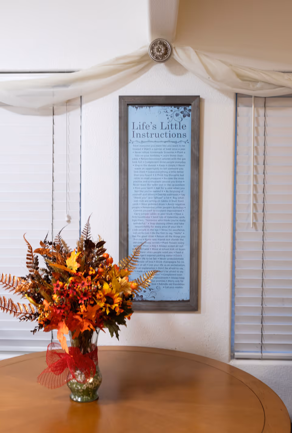 A wooden round table with a glass vase holding an autumn-themed floral arrangement with orange, yellow, and red leaves and flowers. Behind the table is a wall with a framed sign titled 'Life's Little Instructions' and two windows with white blinds. A white sheer fabric is draped above the sign, held by a decorative round wall hook.