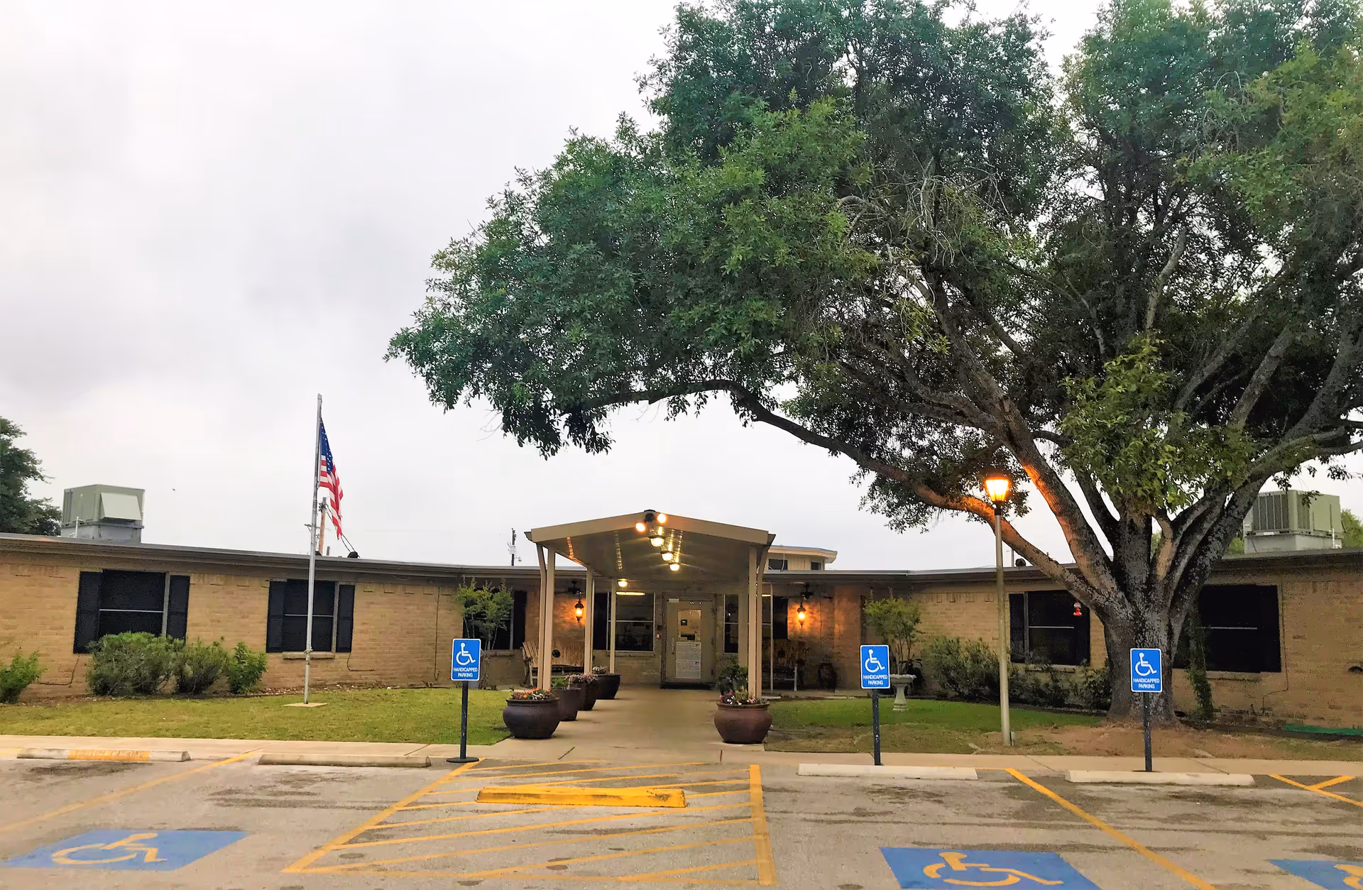 Exterior view of a single-story brick building with a covered entrance, large tree to the right, American flag on a pole to the left, and multiple handicap parking spaces in front.