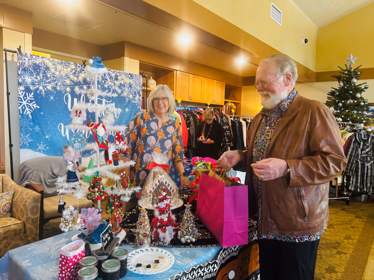 A man and a woman are interacting at a holiday-themed craft sale table inside a senior living facility. The table is decorated with small Christmas trees, ornaments, and festive crafts. Behind them, racks of clothing are visible, and a decorated Christmas tree stands in the background. The woman is smiling and standing behind the table, while the man is holding a pink shopping bag and looking at the crafts. The setting appears warm and inviting with holiday decorations.