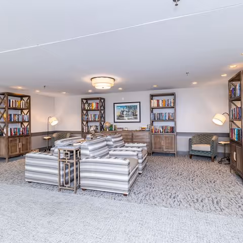A cozy living room area with striped sofas arranged around a small table, flanked by wooden bookshelves filled with books. The room has soft lighting from ceiling fixtures and floor lamps, with additional armchairs placed near the walls. A framed picture hangs on the wall above a wooden cabinet.