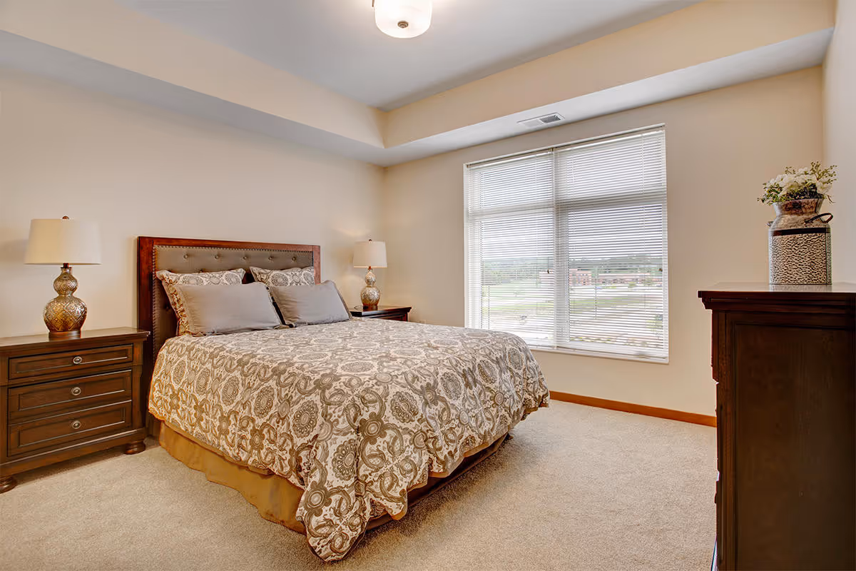Well-lit bedroom with a patterned-bed, two nightstands and lamps, a dresser, and a large window with blinds.