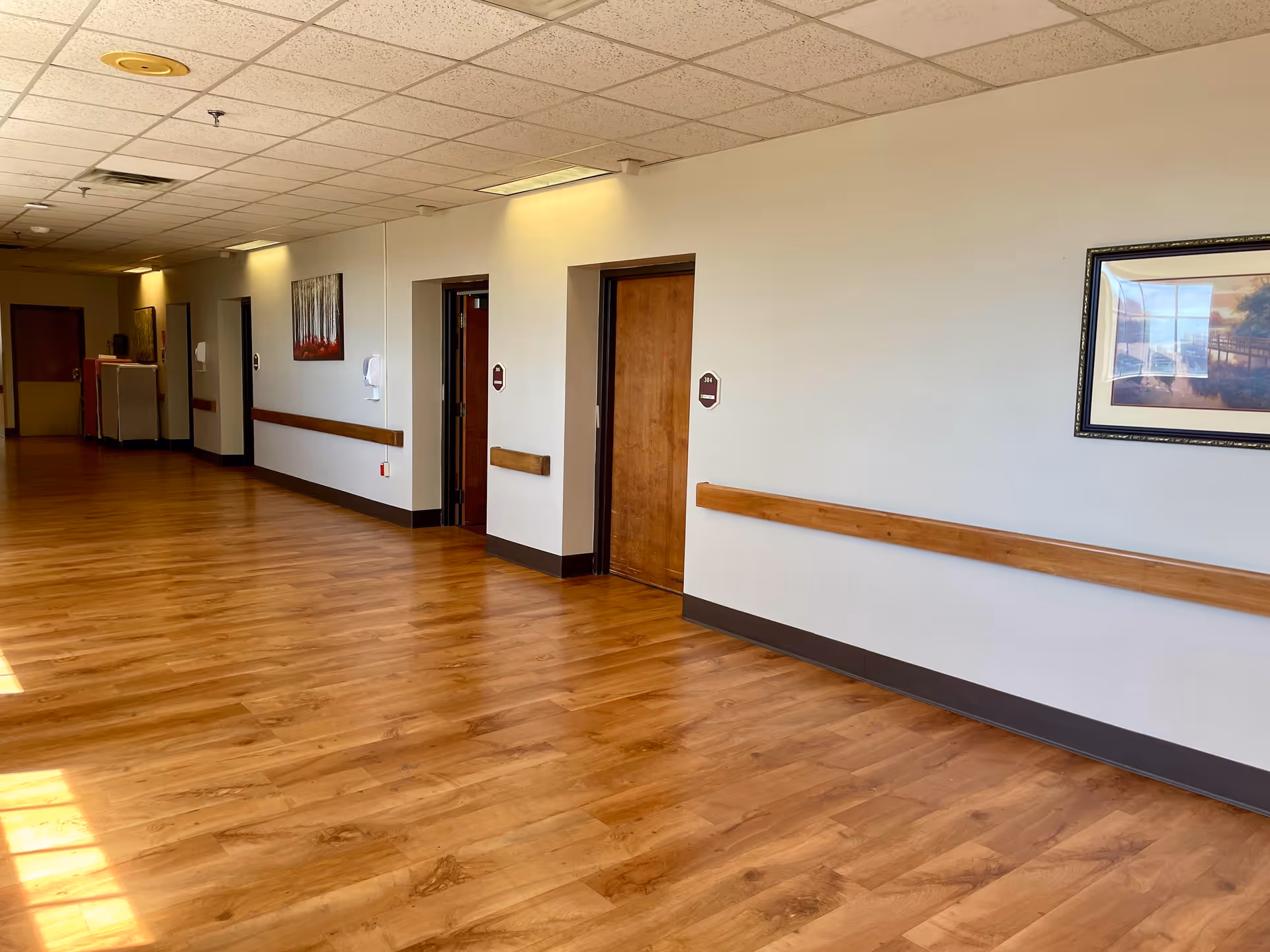 A clean, well-lit hallway in a healthcare facility with wooden flooring, beige walls, and wooden handrails. Several closed wooden doors line the hallway, each with a small sign. There are framed pictures on the walls and ceiling lights illuminating the space.