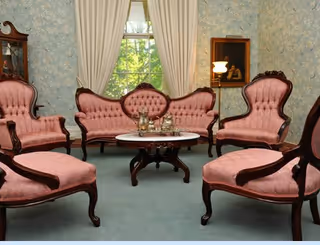 A vintage-style living room with pink upholstered wooden chairs and a matching loveseat arranged around a round wooden coffee table. The room has light blue floral wallpaper, a window with cream curtains, a floor lamp, and a framed painting on the wall.