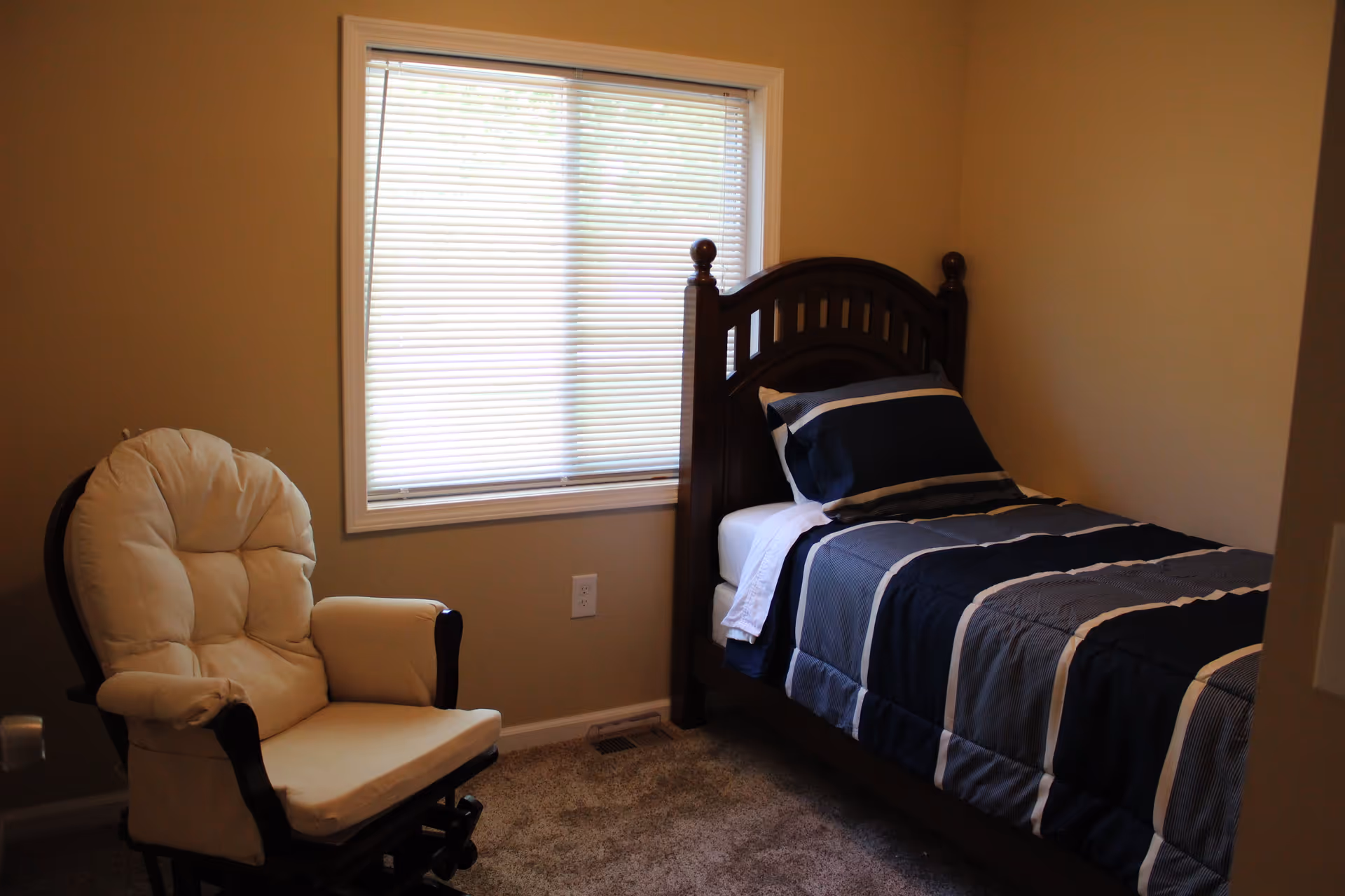 A small bedroom with beige walls and carpeted floor. There is a single wooden bed with a dark blue and gray striped comforter and matching pillow. Next to the bed is a cushioned beige rocking chair. A window with closed white blinds is centered on the wall behind the bed and chair.