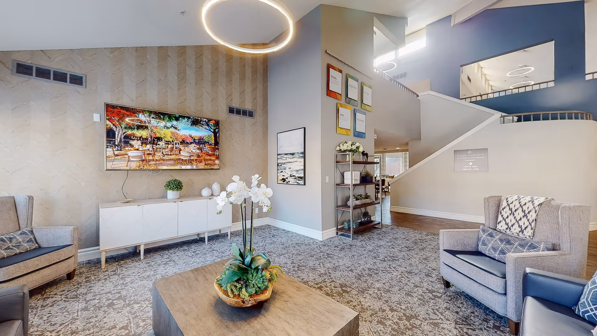A modern living room area with gray armchairs and a wooden coffee table featuring a decorative plant. A large flat-screen TV is mounted on a textured wall above a white console. The room has a patterned carpet, a circular ceiling light, and a staircase leading to an upper floor. There are framed artworks and a shelving unit with plants and decor items.