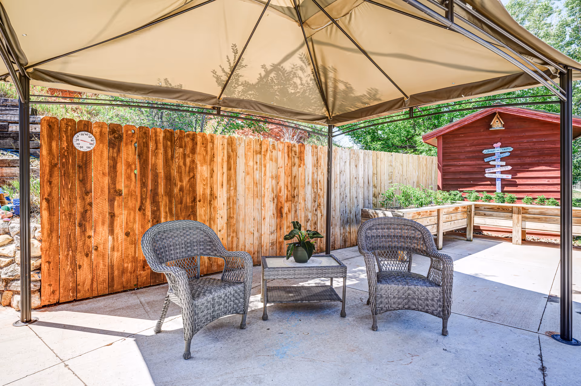 Outdoor patio area with two wicker chairs and a small table with a potted plant under a beige canopy. A wooden fence and a red shed with a directional signpost are visible in the background, along with some greenery.