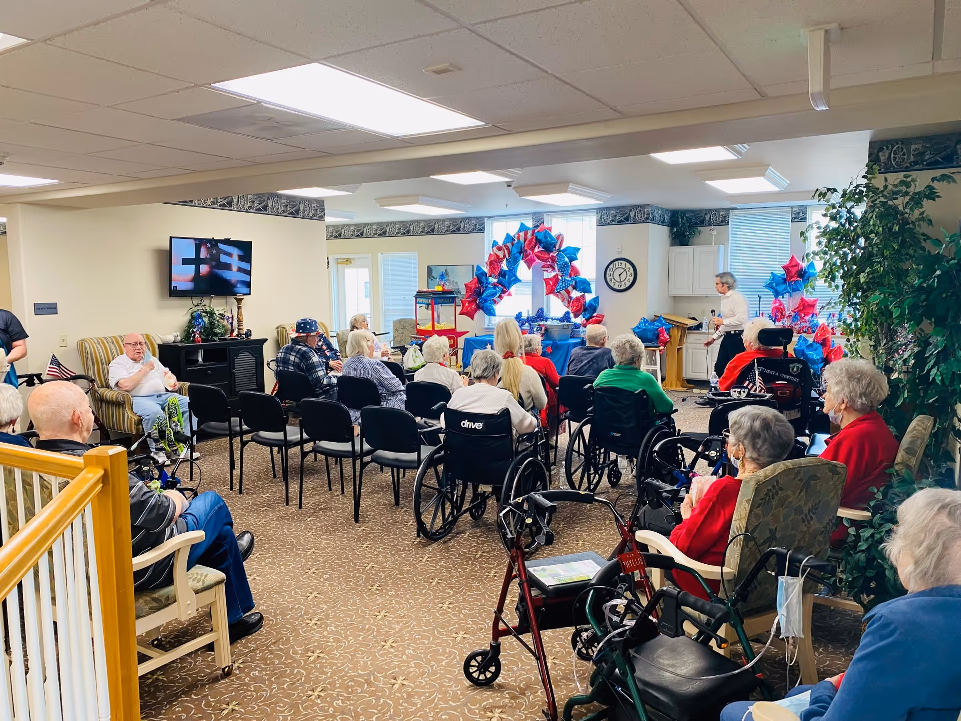 A group of elderly people seated in a common room, some in wheelchairs and others in chairs, facing a woman standing near a podium. The room is decorated with red, white, and blue star-shaped balloons and a patriotic arch. A television mounted on the wall displays an American flag. The setting appears to be a community event or gathering in a senior living facility.