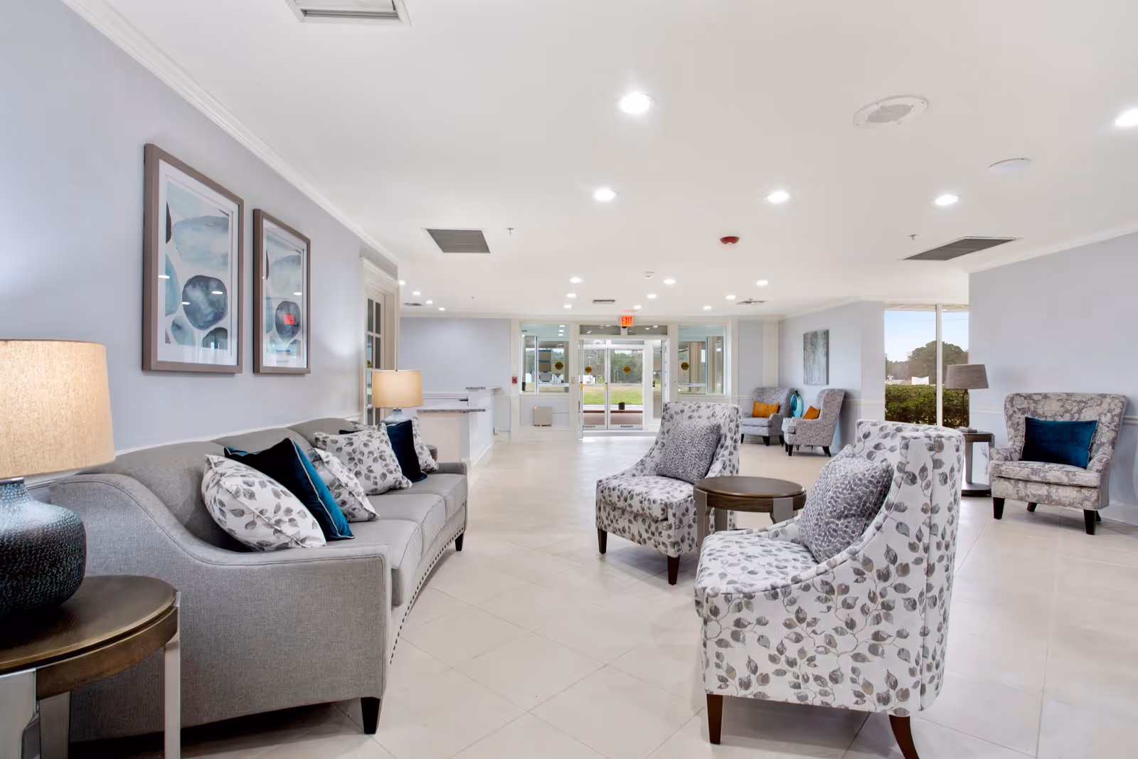 A bright and spacious senior living facility lounge area with a gray sofa adorned with patterned and solid pillows, two patterned armchairs around a small round table, and additional seating near large windows. The room has light-colored tiled floors, soft blue walls, ceiling lights, and framed abstract artwork on the wall.