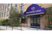 Entrance of Holliswood Center with a purple awning displaying the facility's name and logo. The building exterior is made of brick, with large windows and a ramp leading to the entrance. Some greenery is visible along the side.