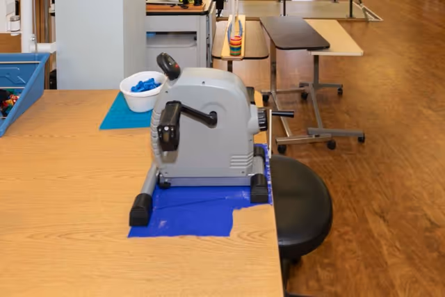 A small gray rehabilitation pedal exerciser sits on a wooden table with a black stool and therapy equipment in a rehab room.