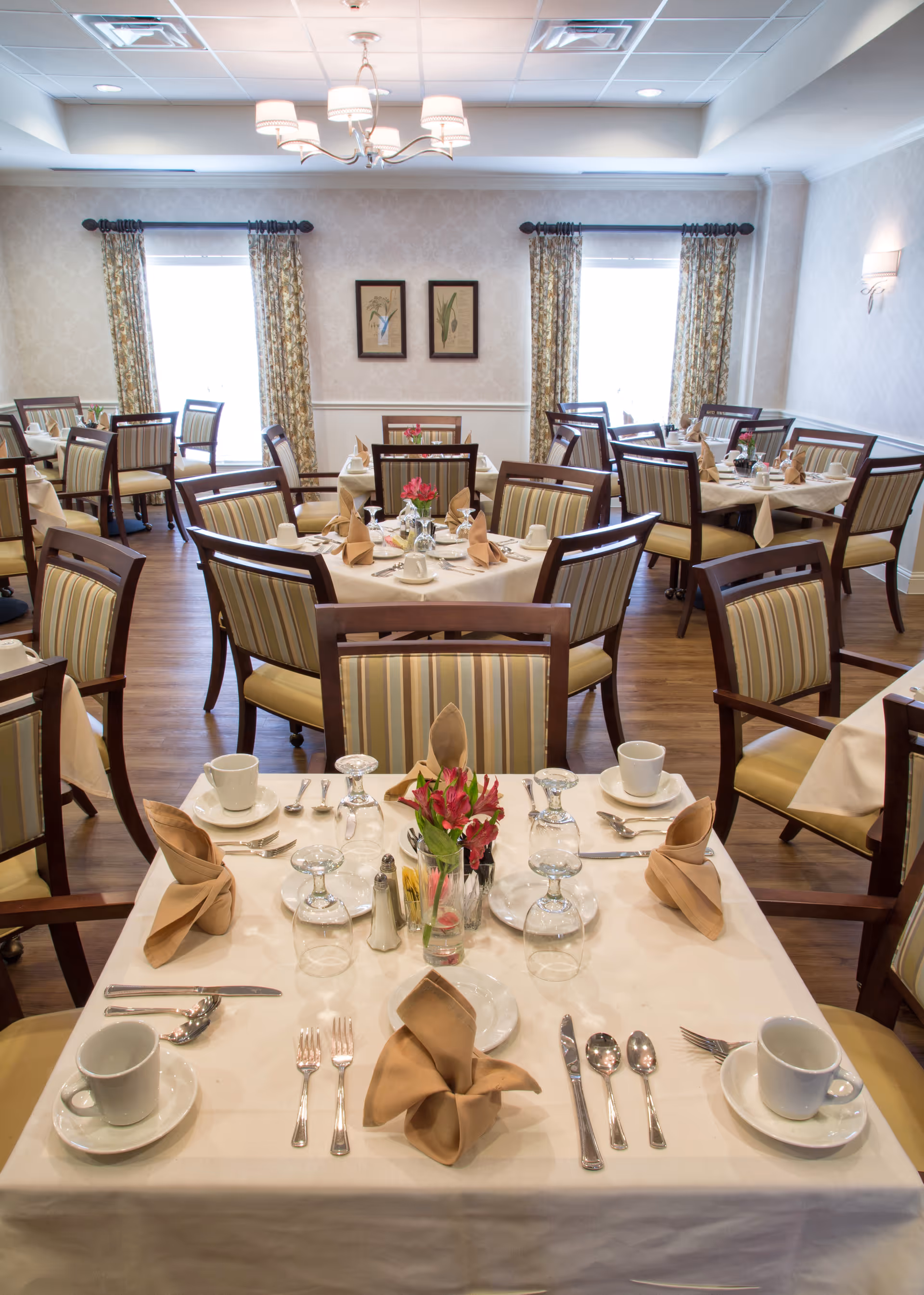 A neatly arranged dining room with multiple tables set for a meal. Each table has beige tablecloths, folded beige napkins, white cups and saucers, silverware, glassware, and small flower arrangements. The room has wooden chairs with striped upholstery, patterned curtains on the windows, and framed botanical artwork on the walls.