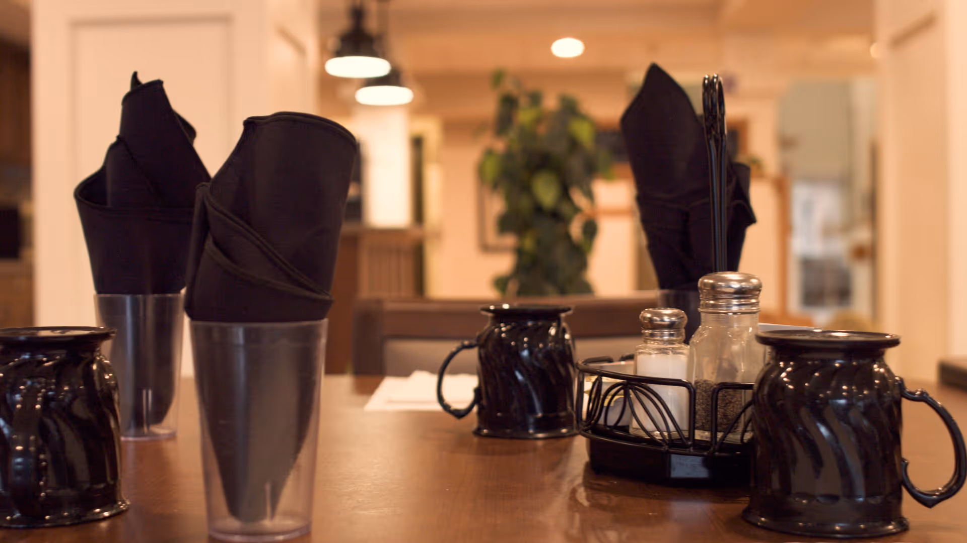 Close-up view of a dining table set with black napkins in clear holders, black ceramic mugs, and a condiment holder with salt and pepper shakers in a warmly lit dining area with blurred background including plants and furniture.