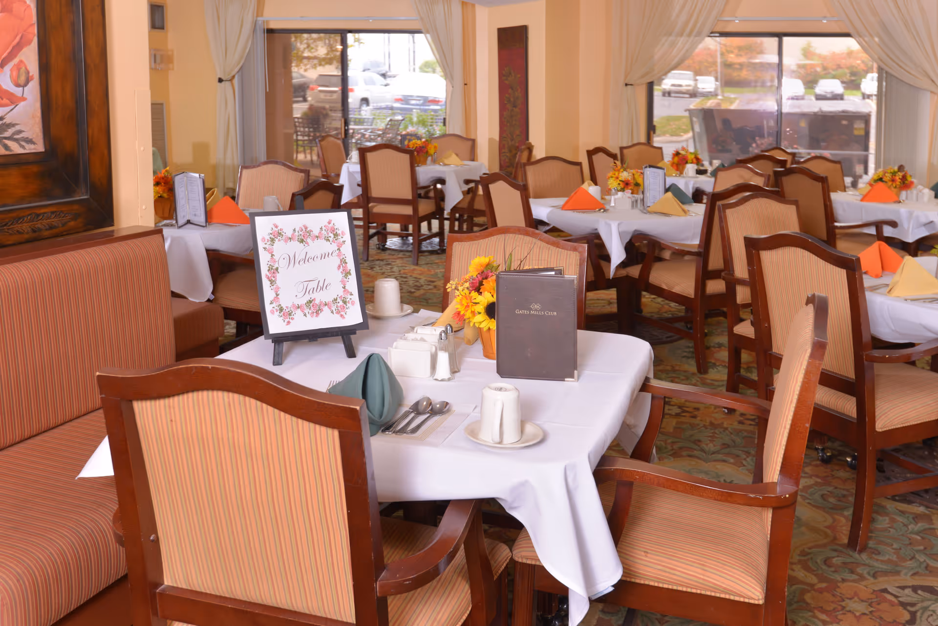 A dining room in a senior living facility with multiple tables covered in white tablecloths, each set with napkins in various colors, menus, and small flower arrangements. There is a sign on one table that reads 'Welcome Table'. The room has large windows with curtains, allowing natural light to enter, and upholstered chairs around the tables.