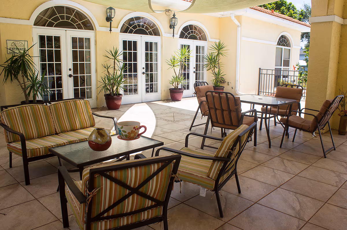 Outdoor patio area at Ashton Place with cushioned chairs and sofas arranged around glass-top tables. The patio is tiled and adjacent to a building with cream-colored walls and white-framed glass doors and windows. Several potted plants are placed near the doors, and sunlight brightens the space.