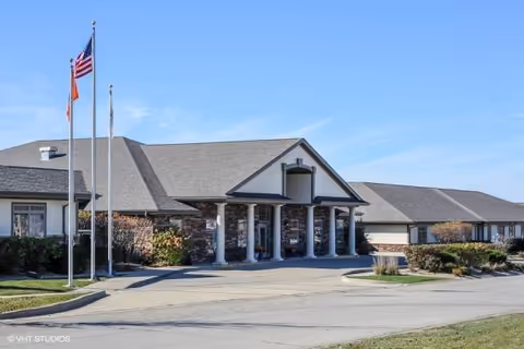 Front exterior of The Glenwood Assisted Living of Mahomet with entrance, flagpoles, and landscaped frontage.