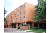 Exterior view of a multi-story brick building with several windows, a covered entrance, and a sign in front surrounded by greenery and trees under a clear sky.