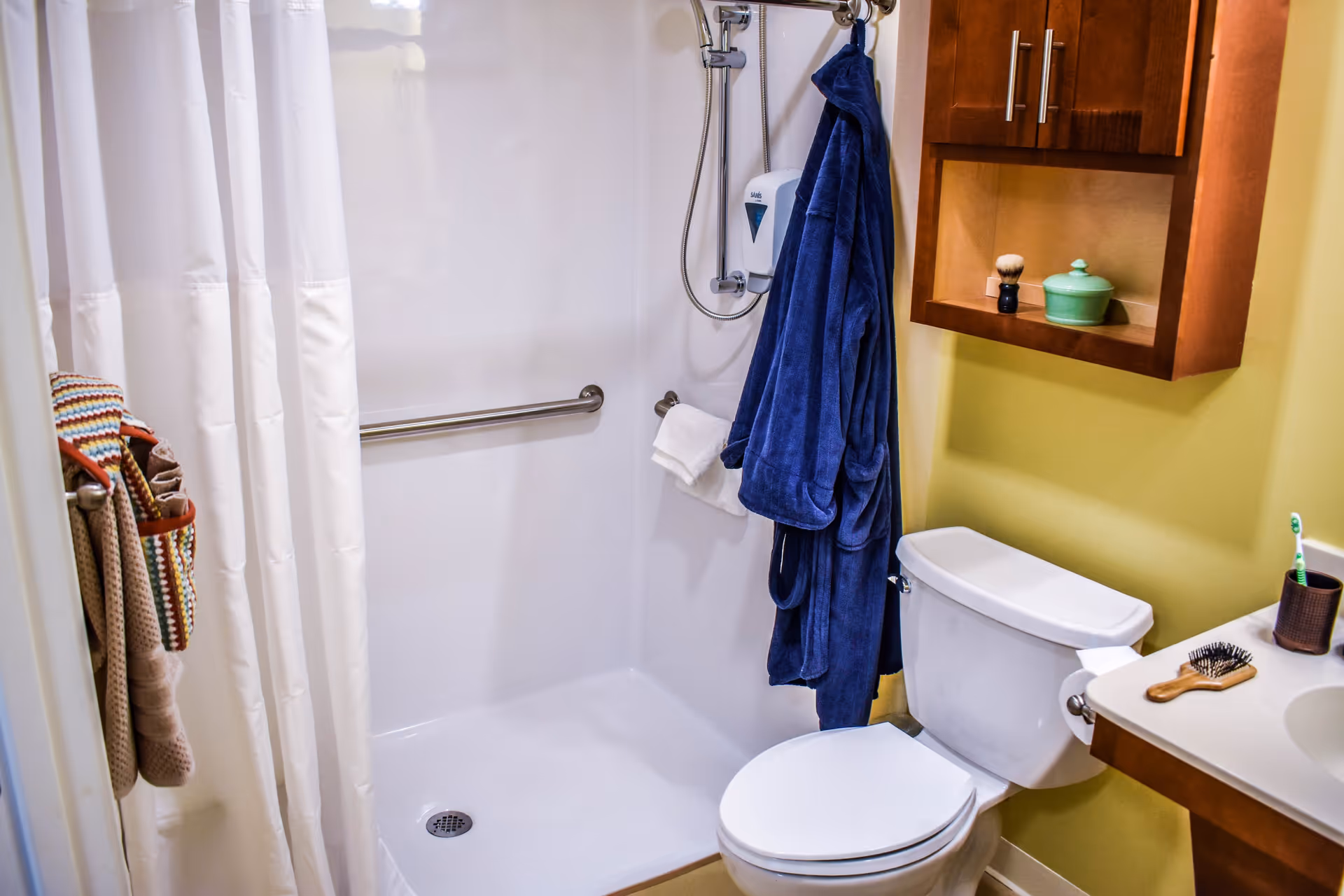 A bathroom with a white shower area featuring a grab bar and a mounted soap dispenser. A blue bathrobe hangs on a hook next to the shower. There is a white toilet beside the shower and a wooden cabinet mounted on the yellow wall above it. On the right, a sink countertop holds a hairbrush and a toothbrush in a holder. Towels hang on a rack near the shower curtain.