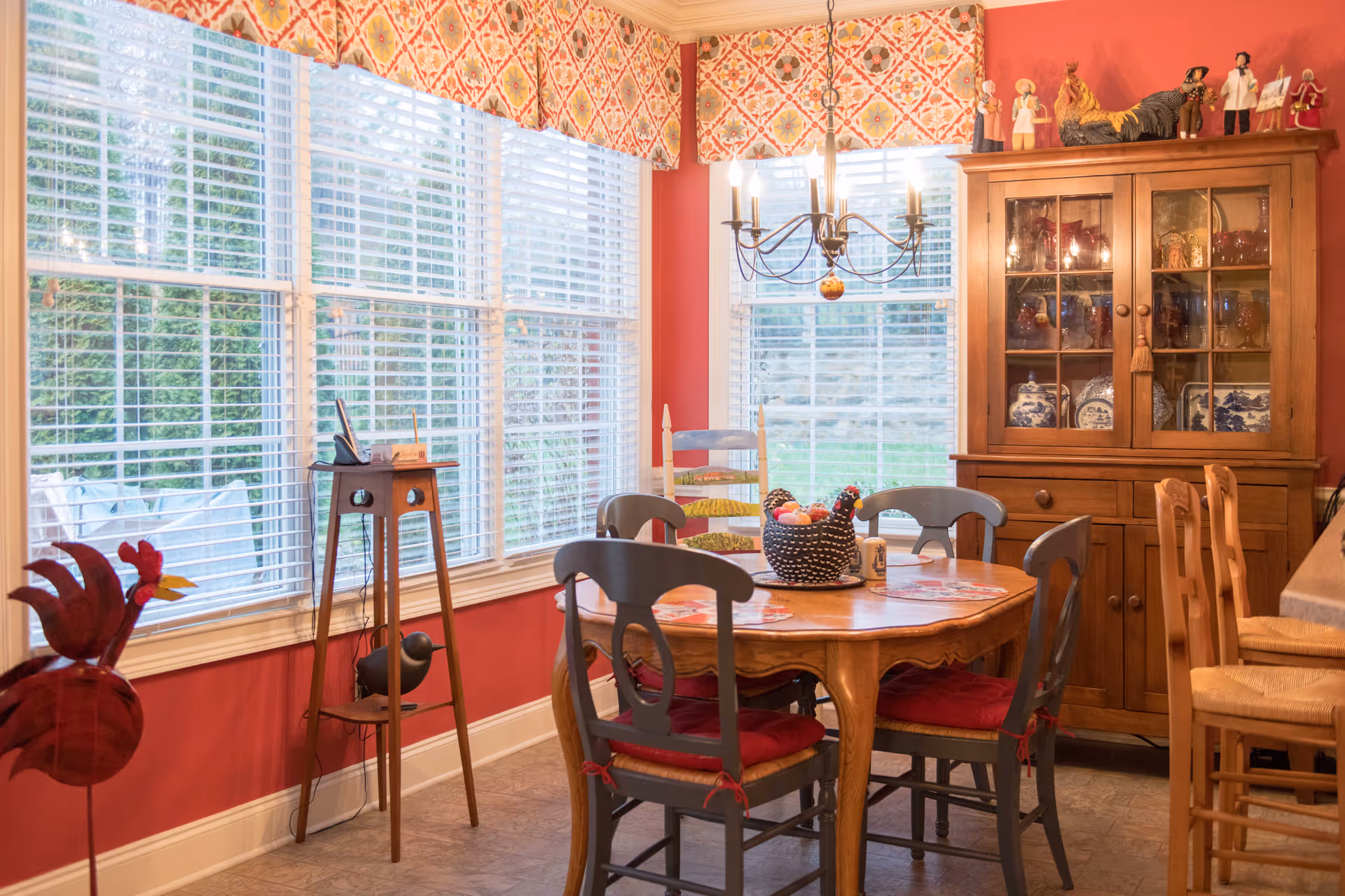 A cozy dining room with red walls and large windows covered with white blinds and patterned valances. The room features a wooden dining table with four chairs, each with red cushions, and a decorative centerpiece shaped like a chicken. A wooden cabinet with glass doors displaying dishes and figurines stands against the wall. A chandelier with lit candles hangs above the table.