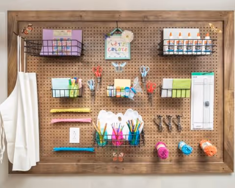 A wooden framed pegboard mounted on a wall, organized with various craft supplies including scissors, glue sticks, colored pencils, markers, paper, and aprons hanging on the left side. The pegboard has multiple wire baskets holding the supplies and a small sign in the center that says 'Let's Create'.