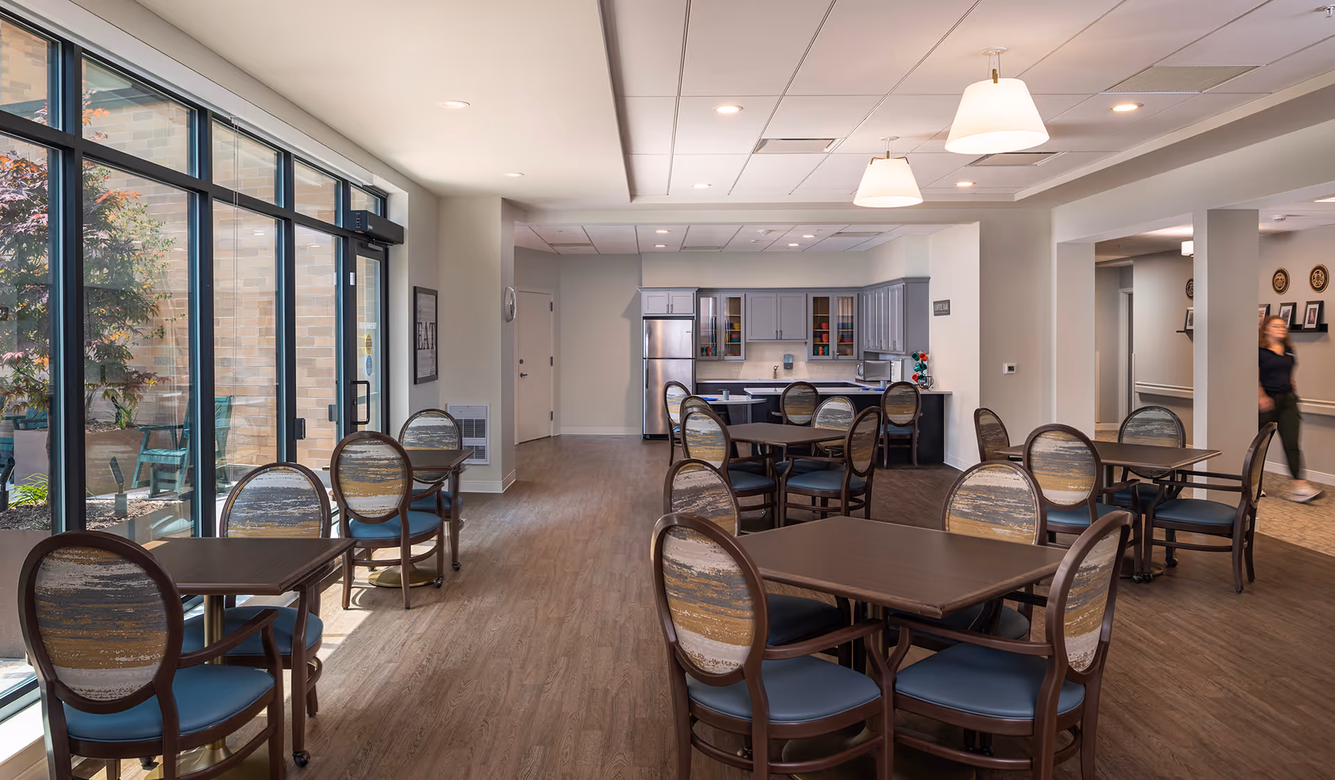 A bright dining area in a senior living facility with several tables and chairs arranged neatly. Large floor-to-ceiling windows on the left side let in natural light and offer a view of an outdoor patio with greenery. At the back, there is a kitchen area with gray cabinets, a refrigerator, and a microwave. A person is walking in the hallway on the right side of the image.
