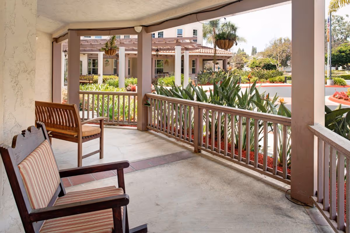 Covered outdoor patio area with wooden chairs and a bench, overlooking a garden with green plants and hanging flower pots. The background shows part of a building and a clear sky.